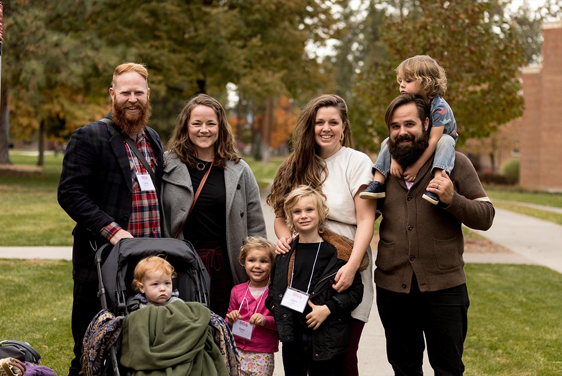 Two young families stand outside, smiling together. One child is in a stroller.