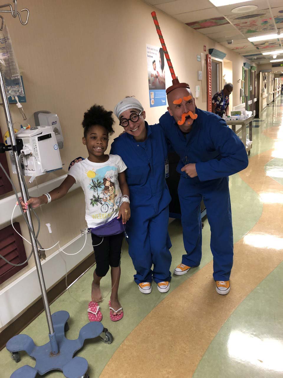 Alex Smith and another medical professional, both in silly costume pieces, smiles with a child in a hospital ward.