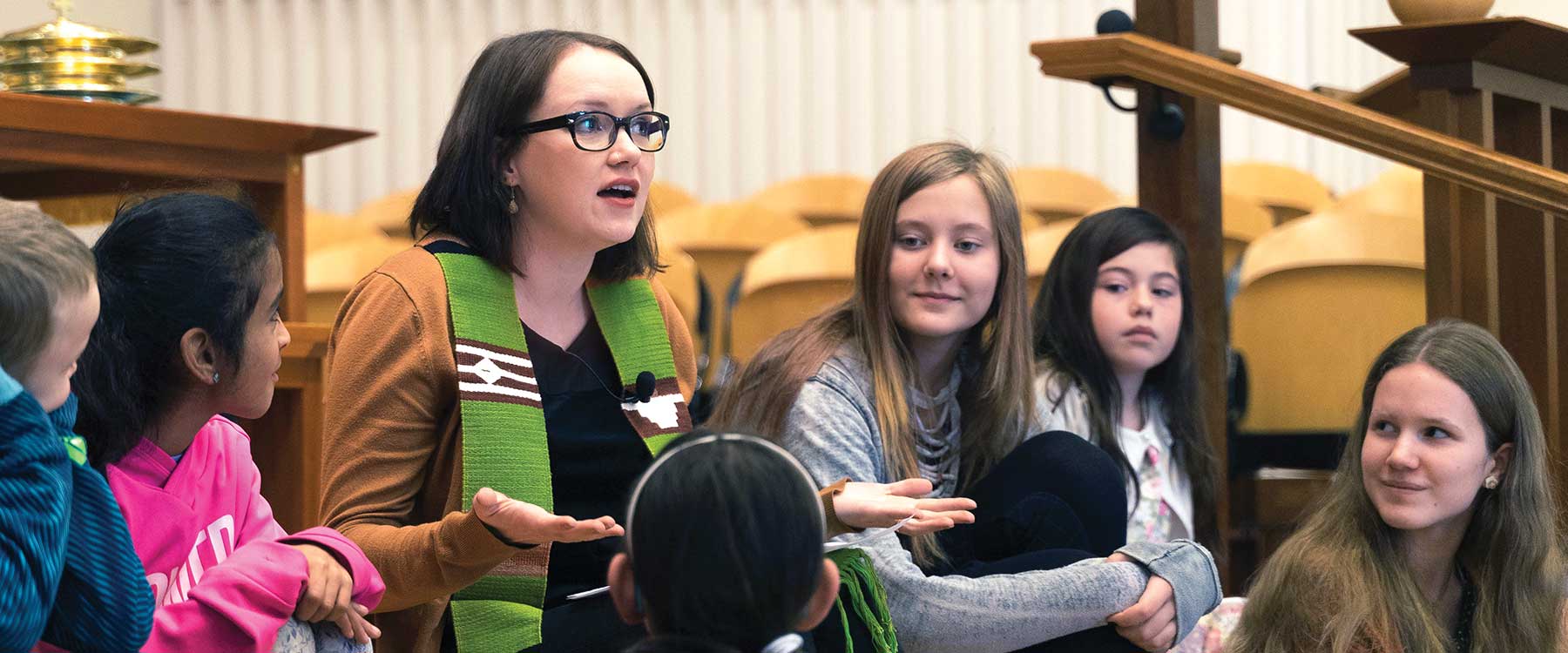 A speaker addresses a group of children, sitting with them on the steps of a chapel stage.