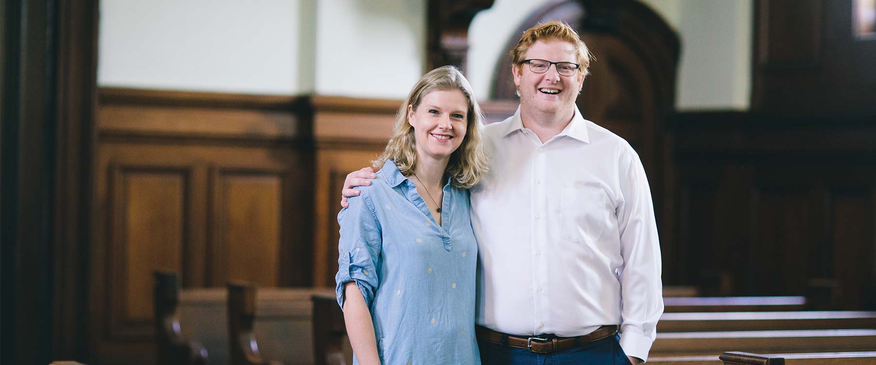 Chris and Jen Atwood stand among the pews of their church. Chris puts his arm on Jenn's shoulder, they smile.