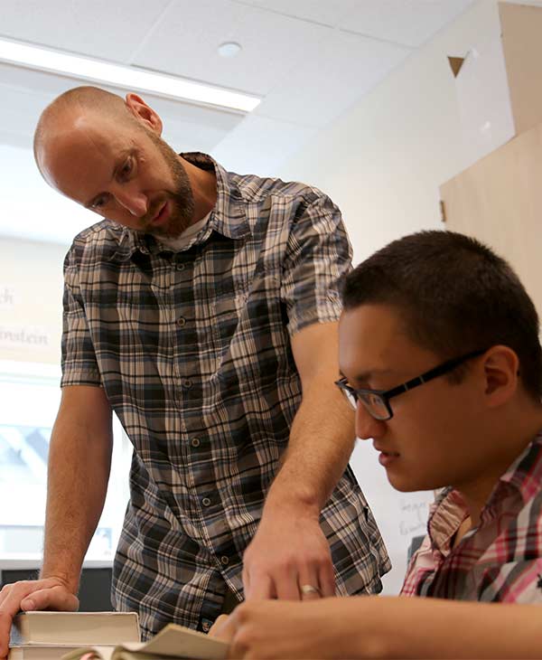 Grant stands next to a seated student. He points to an open book as he talks with the student.