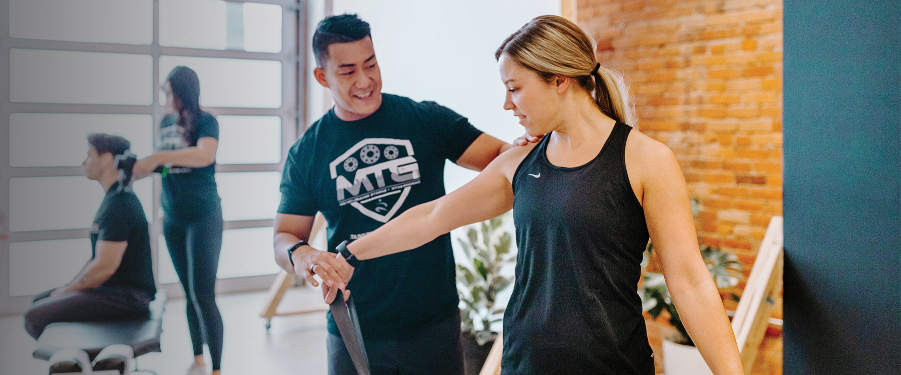 A fitness instructor guides a client in exercise, while another trainer assists a seated person in the background.