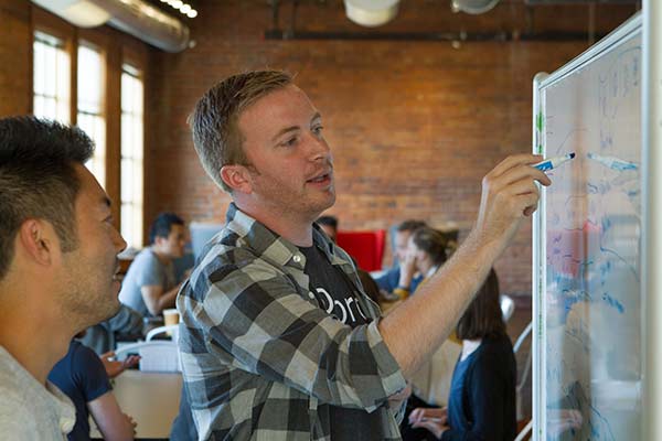 Jake writes on a whiteboard as a co-worker watches.