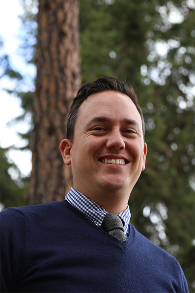 John Pell stands in front of a pine tree on campus and smiles.