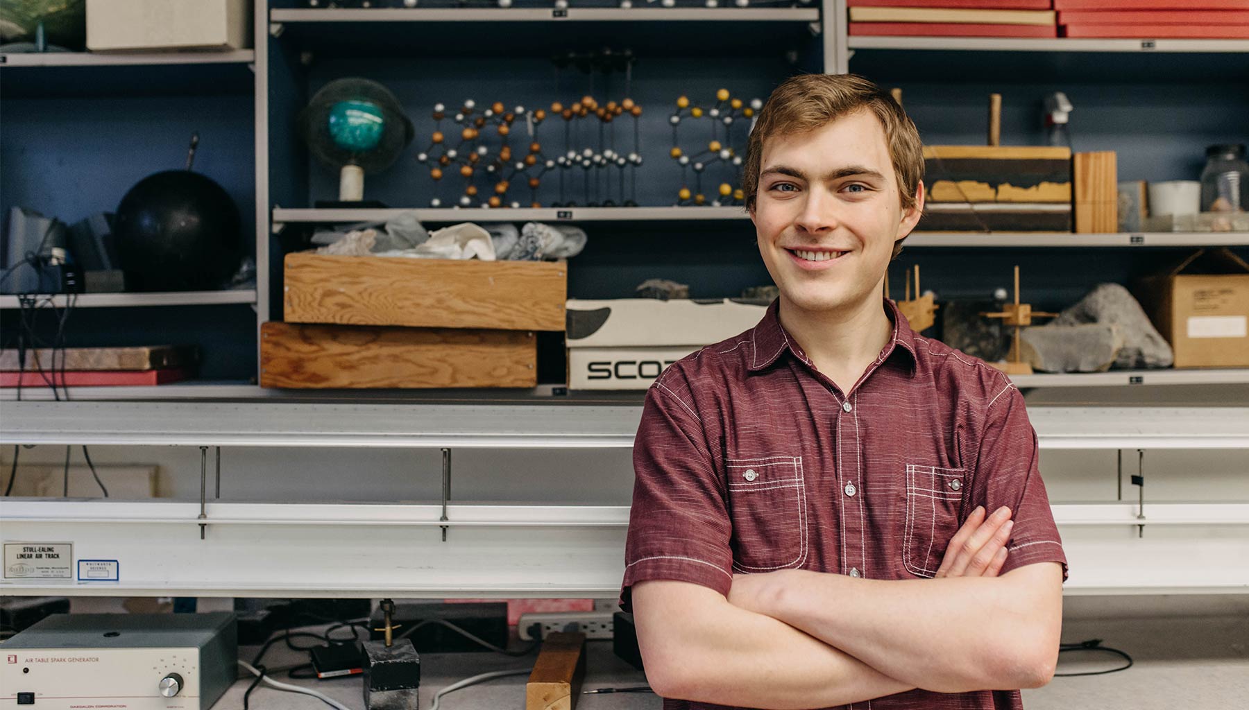 Jorin Graham stands in front of shelves filled with physics models. He folds his arms and smiles.