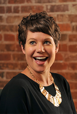 Kendel stands in front of a brick wall and smiles a wide surprised smile.