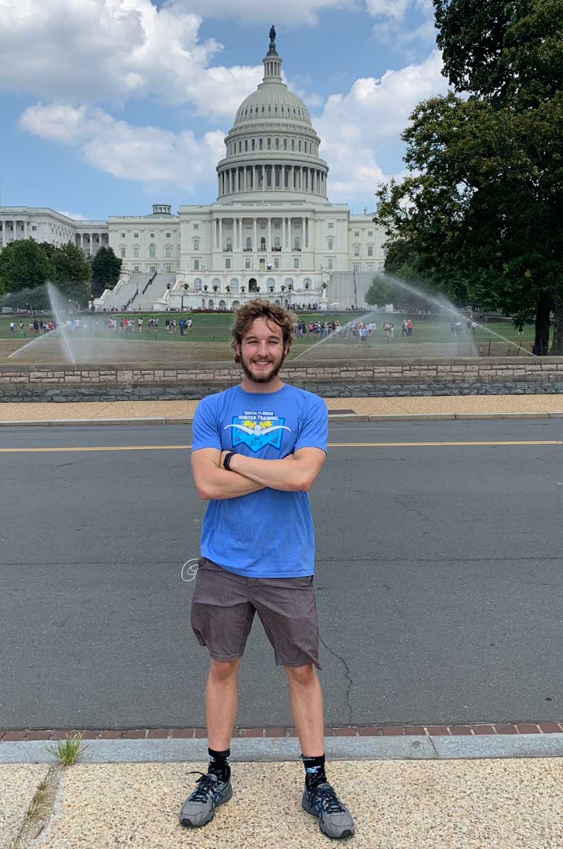 Cannon Coats stands with arms crossed in front of the U.S. Capitol building on a sunny day.