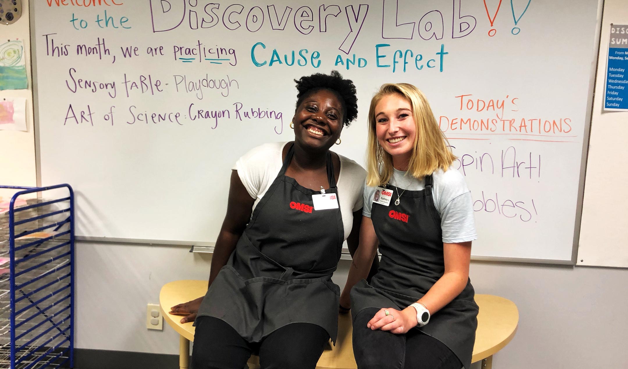 Two smiling people in aprons sit on a table in front of a whiteboard labeled 