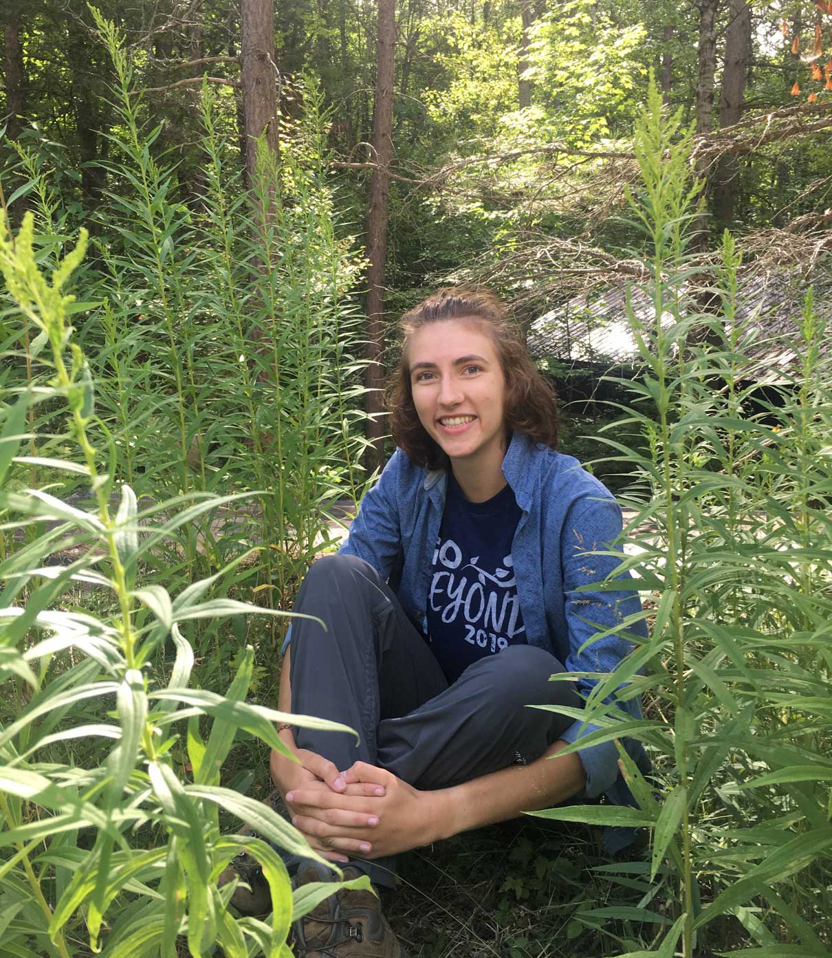 Emily Donnel sits in a forested area and smiles at the camera.