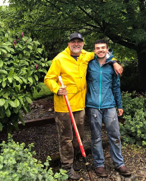 Two people in rain jackets smiling and posing in a garden; one is holding a shovel.