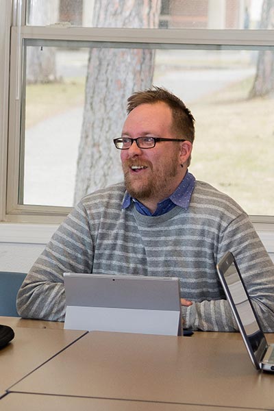 Thom Caraway sits behind a tablet computer in a classroom. He smiles as he talks.