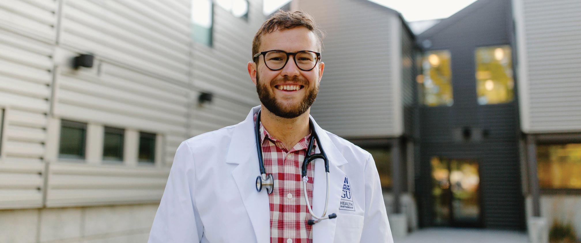 Kevin Glover, wearing a stethoscope and a white coat, stands smiling in front of a building.