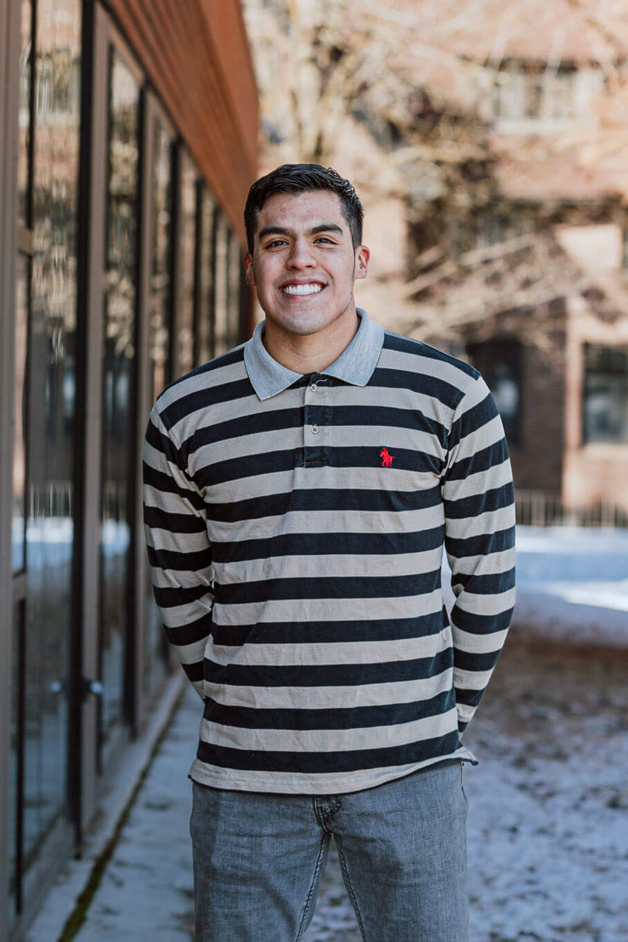 Smiling person standing outdoors in front of a brick building, wearing a black and gray striped long-sleeve shirt.