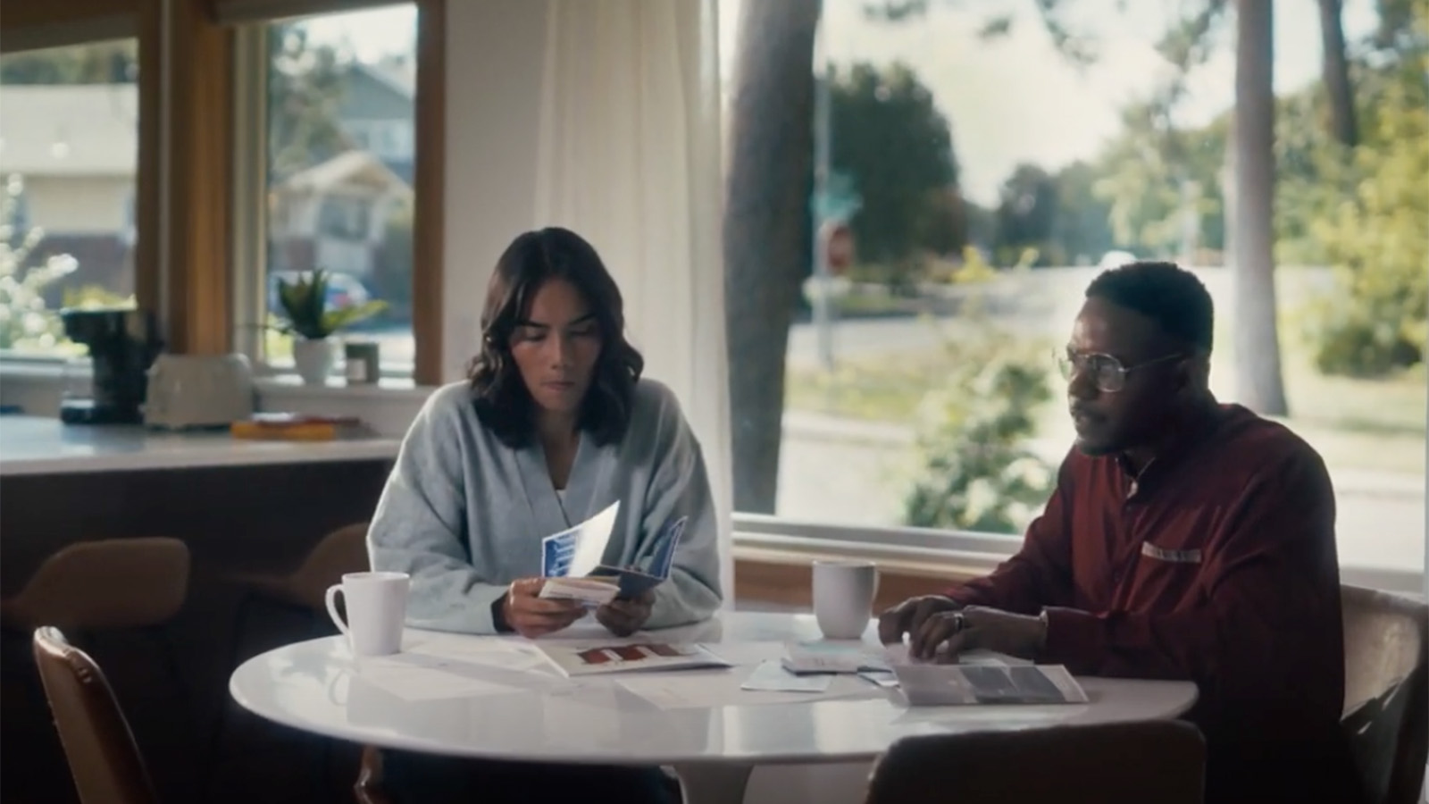 Two people sitting at a round table in a bright kitchen, looking over papers and brochures with coffee mugs in front of them.