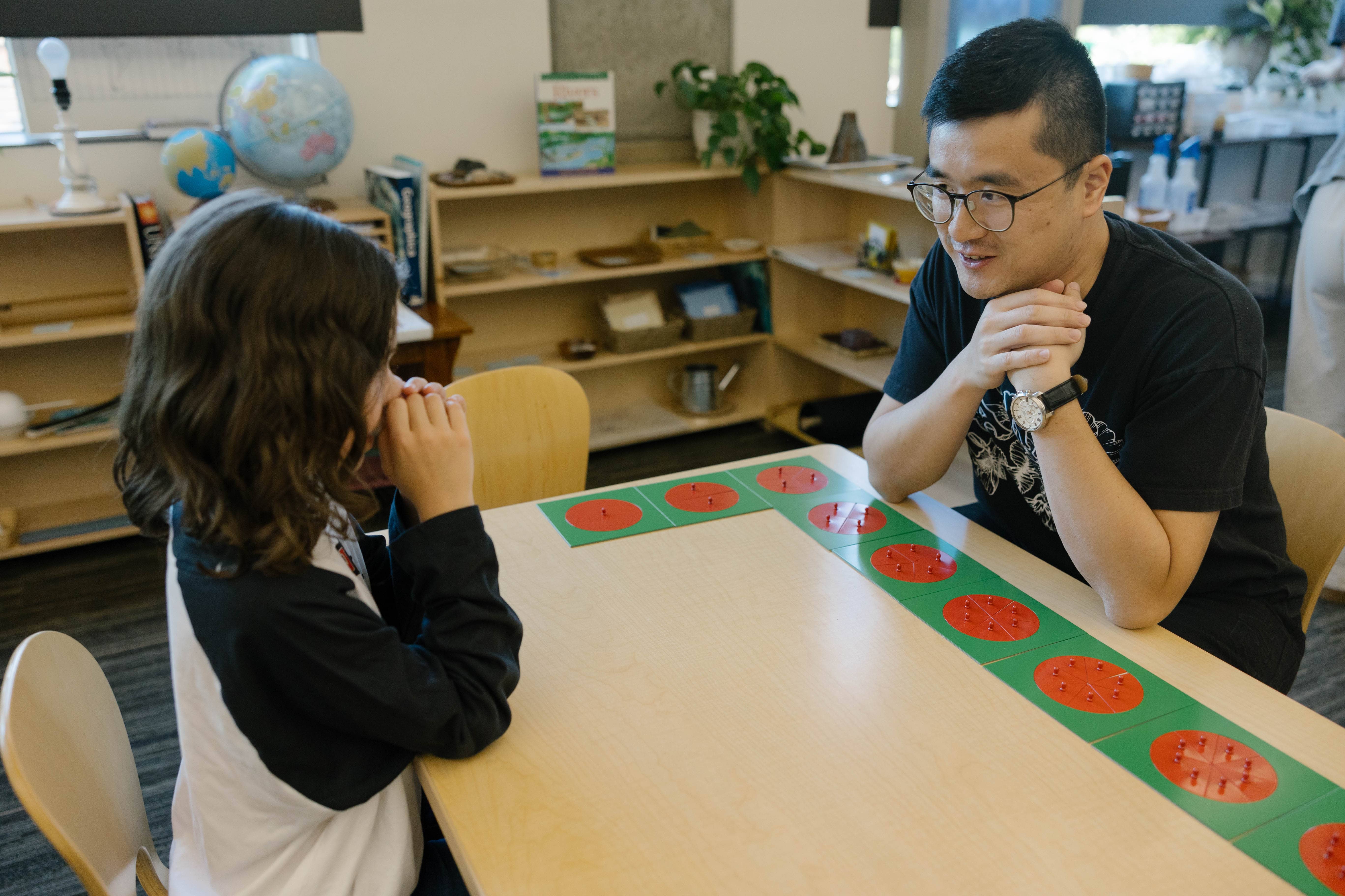An instructor and a young student sit at a small desk discussing a set of fractions toys.