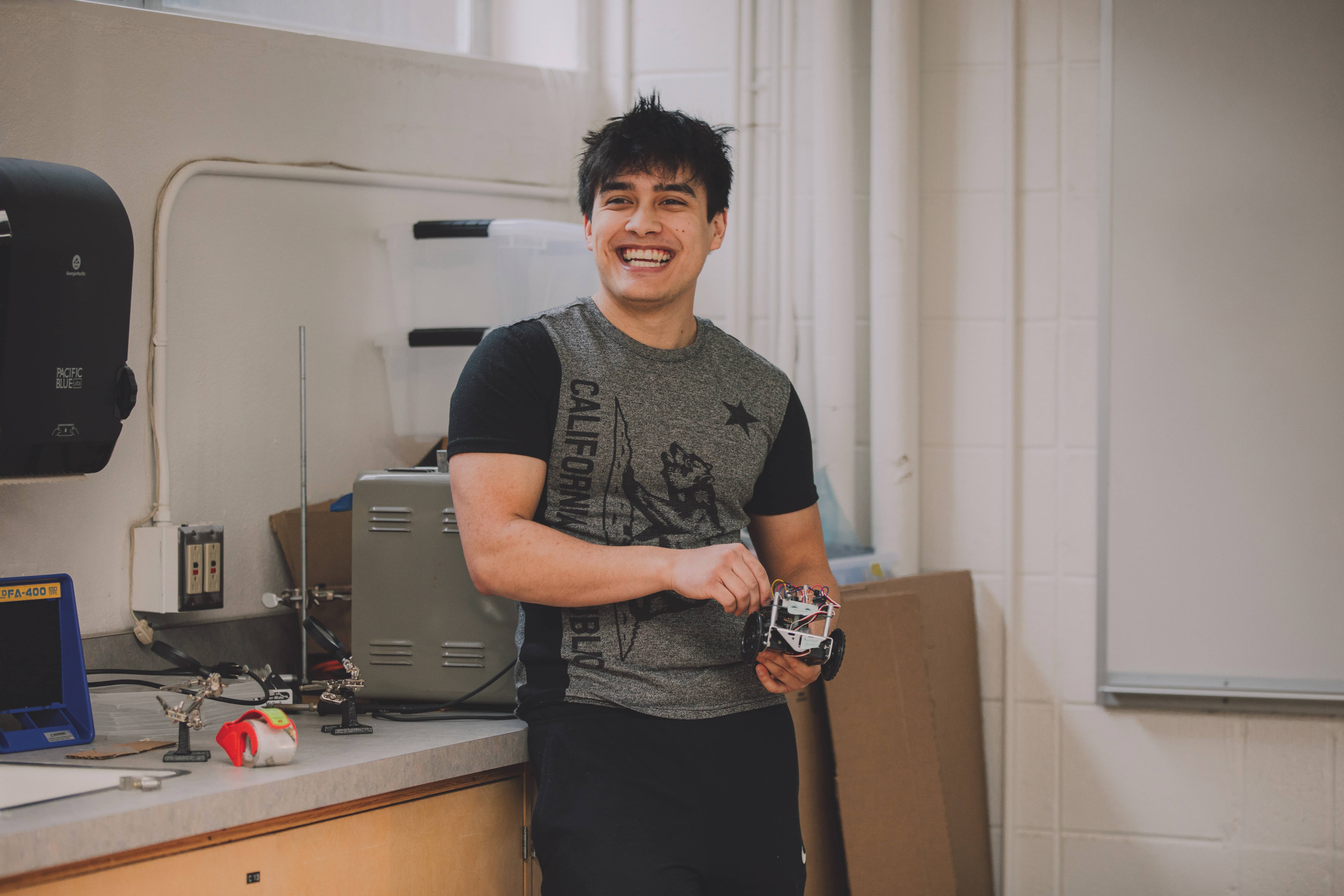 A student stands, smiling, in a lab, holding a small electronic device.