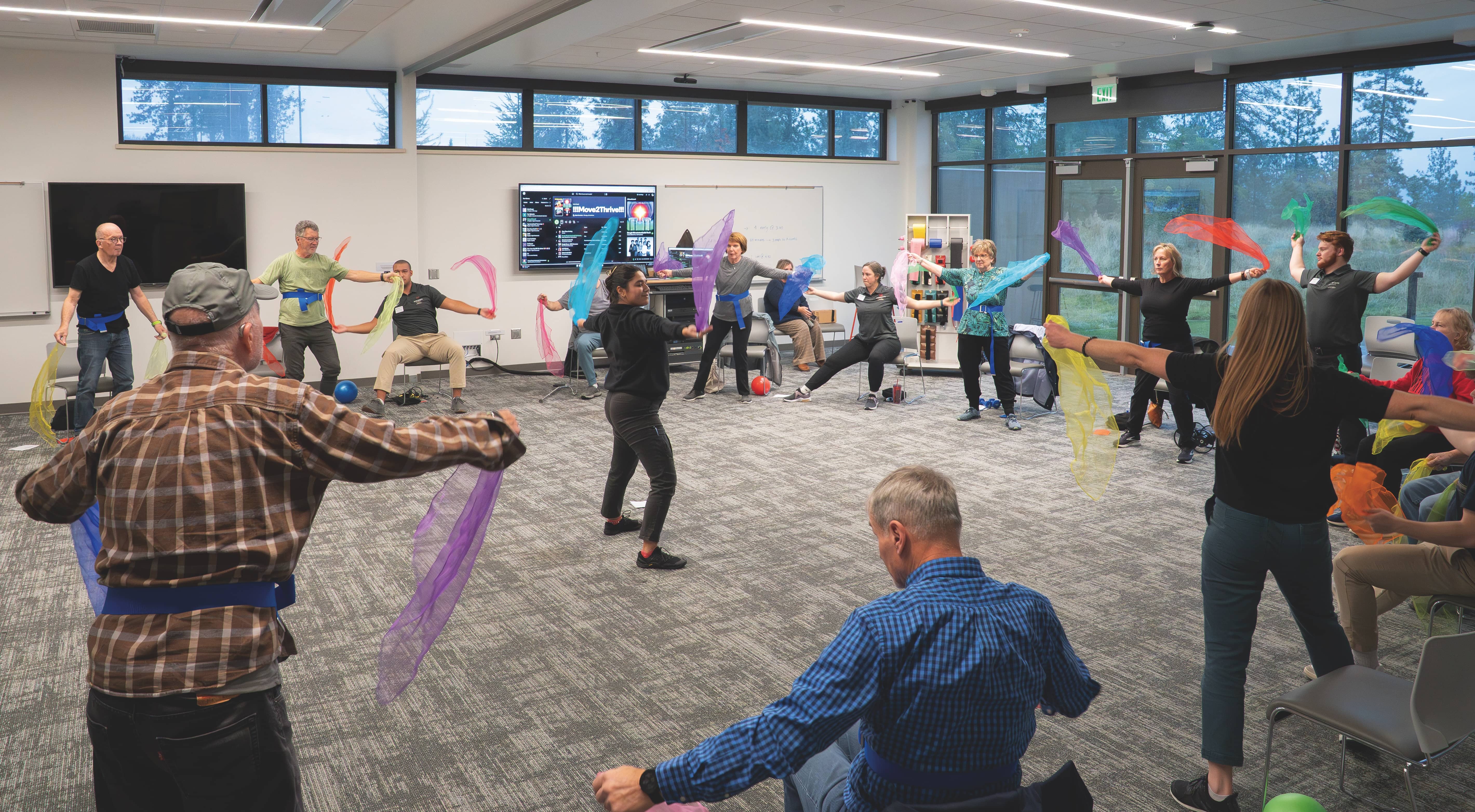 A roomfull of men and women stand and sit in a circle, following the actions of an exercise instructor.