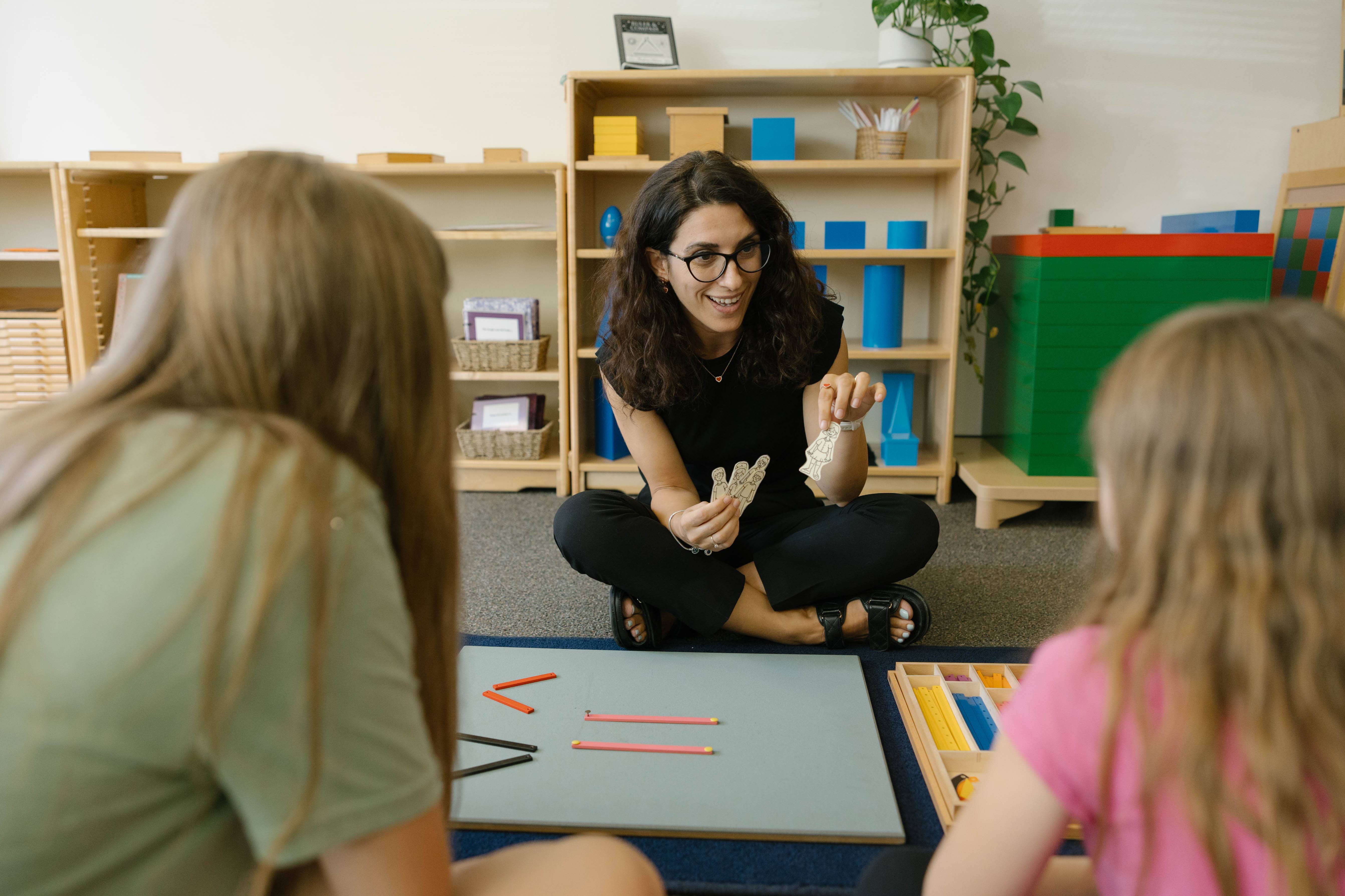 An instructor sits on the floor, facing two young students, and displays paper dolls.
