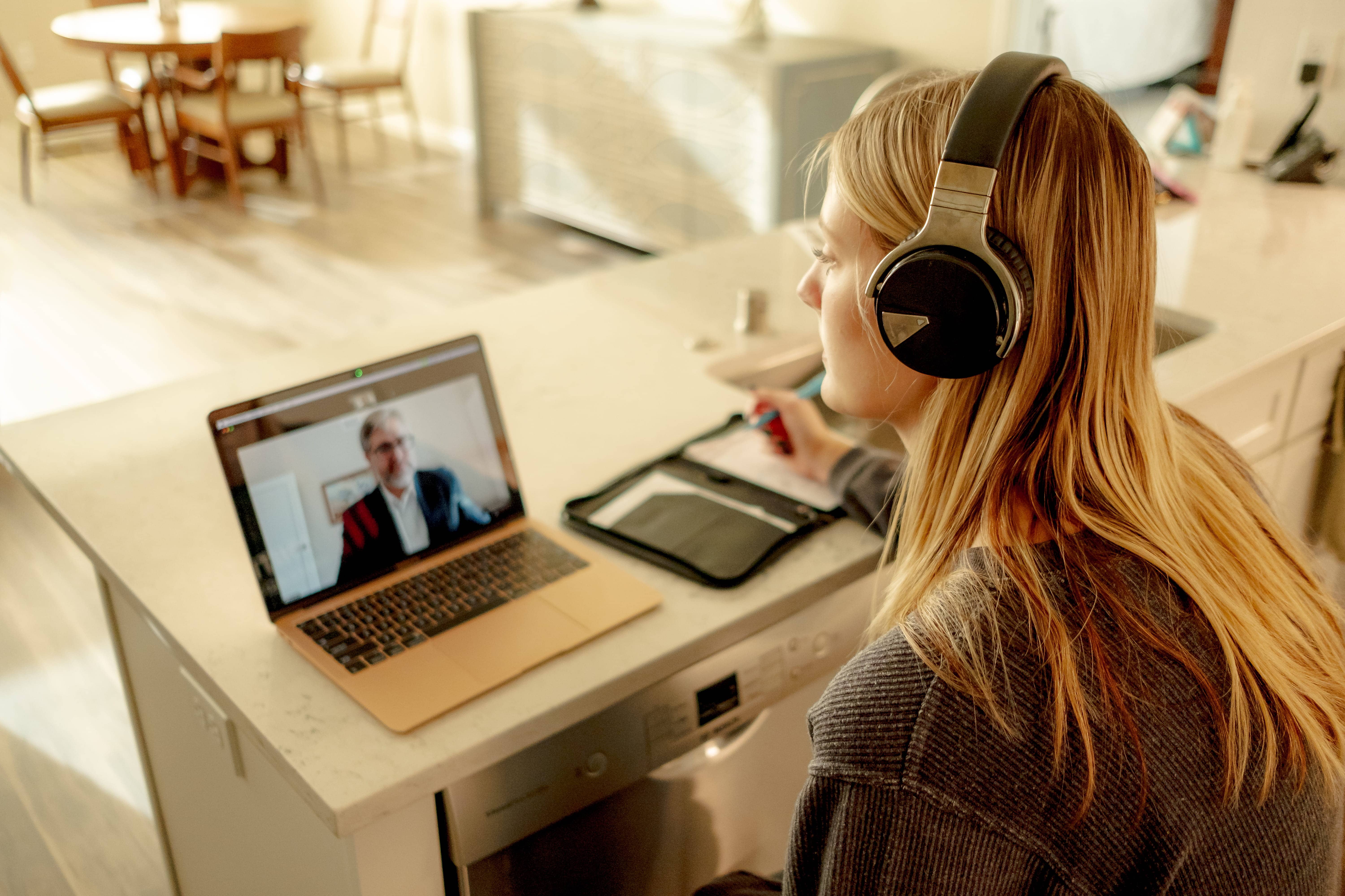 An adult student sits at a kitchen table participating in an online meeting with their instructor.