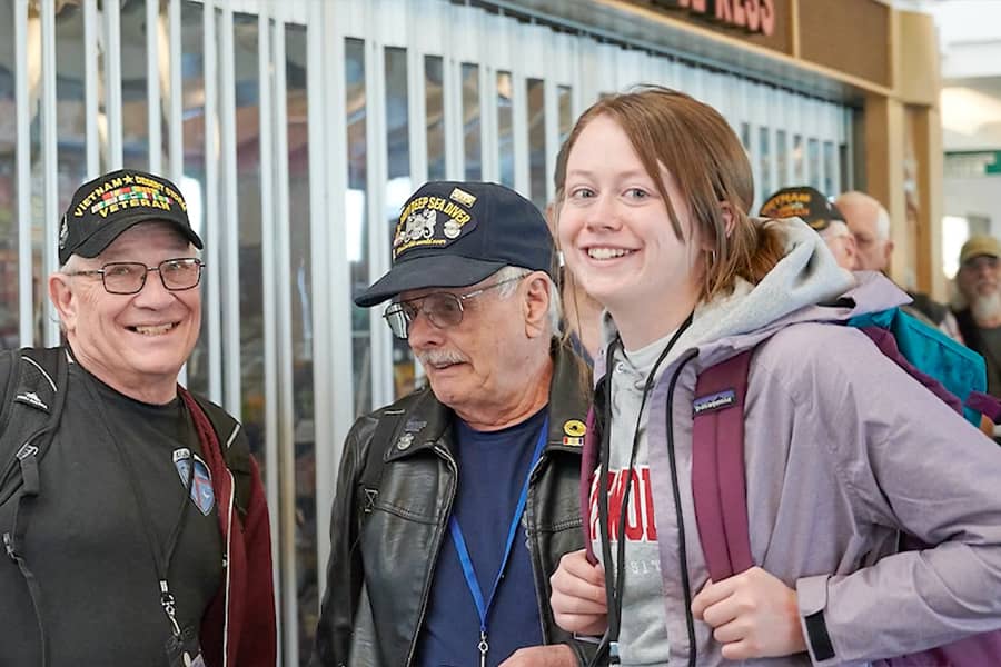 One student and multiple elderly veterans fill an interior hallways, conversing and smiling.