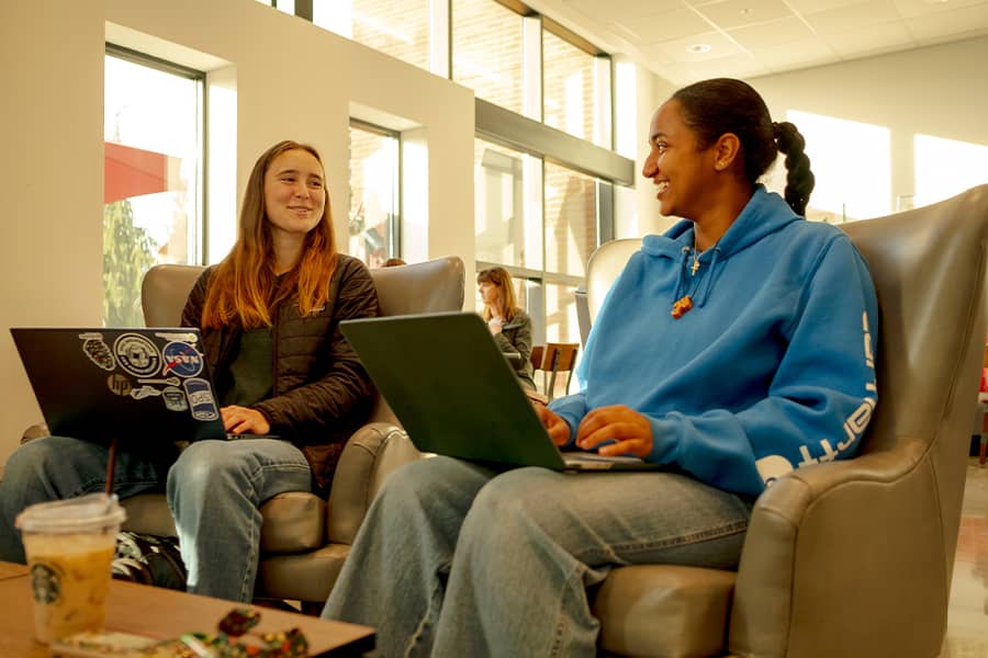 Two students sit in a spacious cafe with computers, chatting.