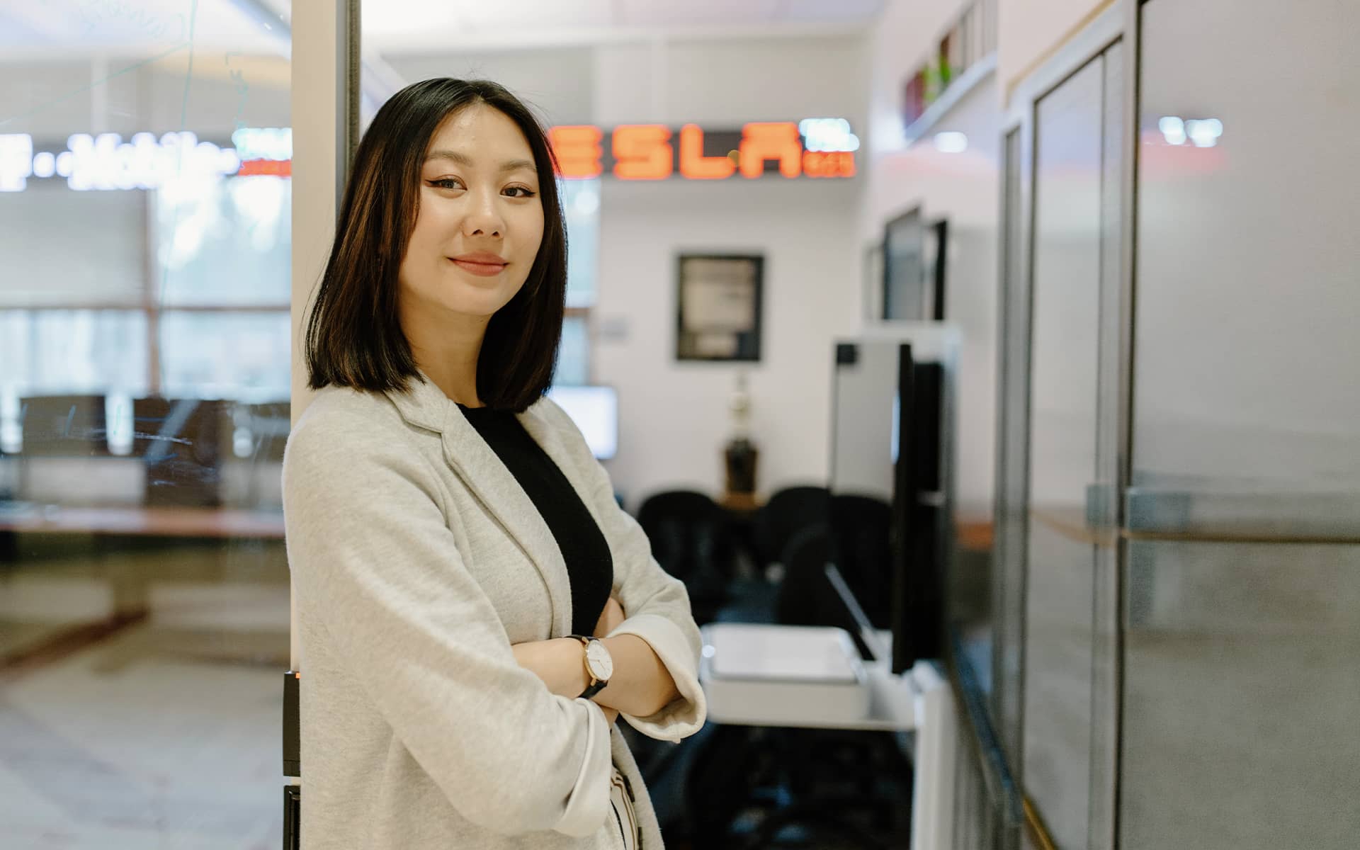 A young professional stands, arms cross and smile, in a stock trading room.