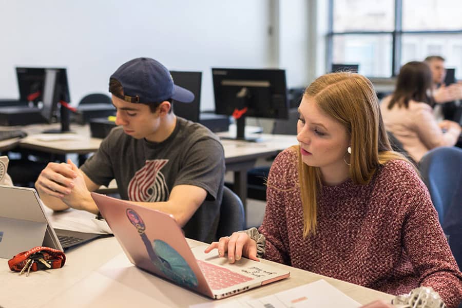 Two students share a desk in a classroom setting, working on laptops.