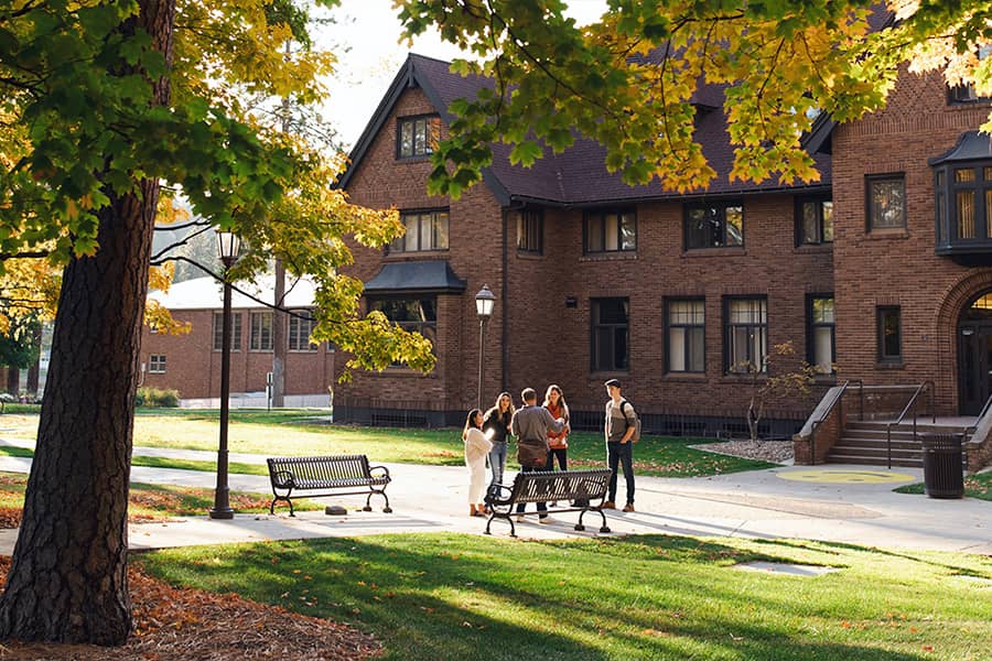 Multiple students stand outside an academic building on a fall day, conversing.