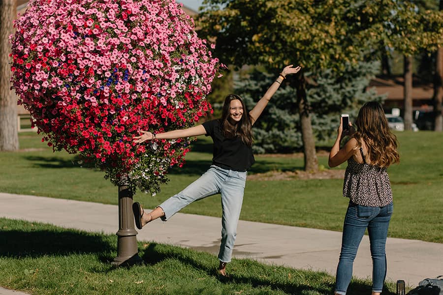 One student poses in a fun gesture in front of a hanging basket of petunias while another takes a photograph.