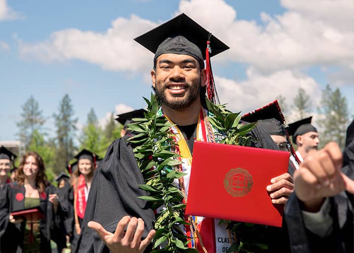 A graduate stands in the forefront, with other graduates behind, wearing a garland and smiling.