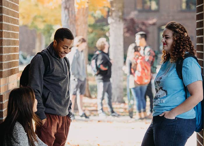 Three students stand around the entryway to an academic building, conversing pleasantly.