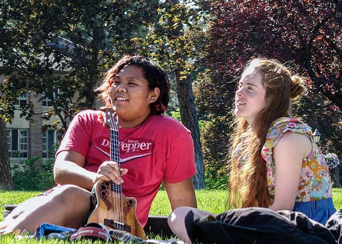 Two students sit on a sunny lawn, smiling, one holds a tenor guitar.