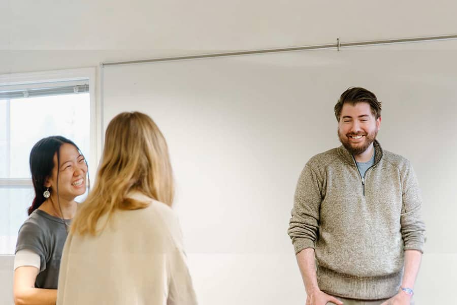 Three students converse and laugh inside a classroom next to a whiteboard.