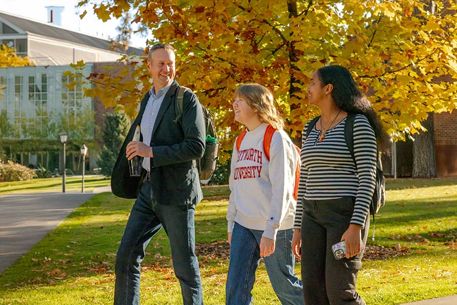 A professor and two students walk outside on a sunny day past an academic building.