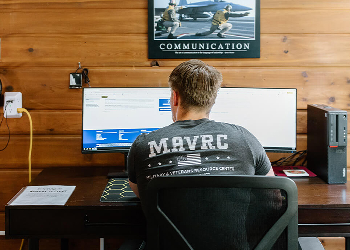 A student wearing a MAVRC T-shirt sits at a desk and works on dual monitors.