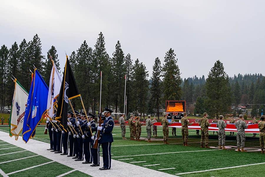 Military service members for two groups, one stands at attention while the other unfurls a large flag.