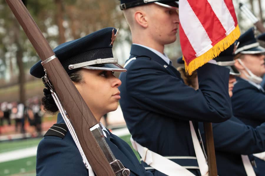 A row of military personnel stand at attention with flags and rifles.