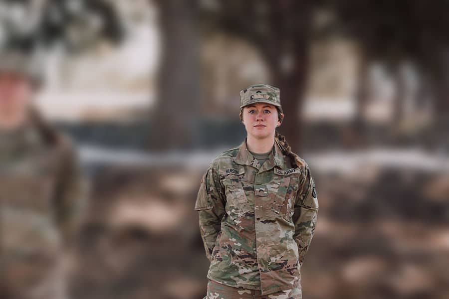A military student in camouflage gear stands with a serious expression. The background is blurred.