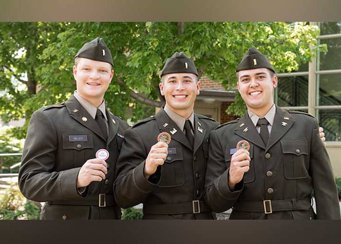 Three military students pose in full military dress, smiling and holding up medalions.