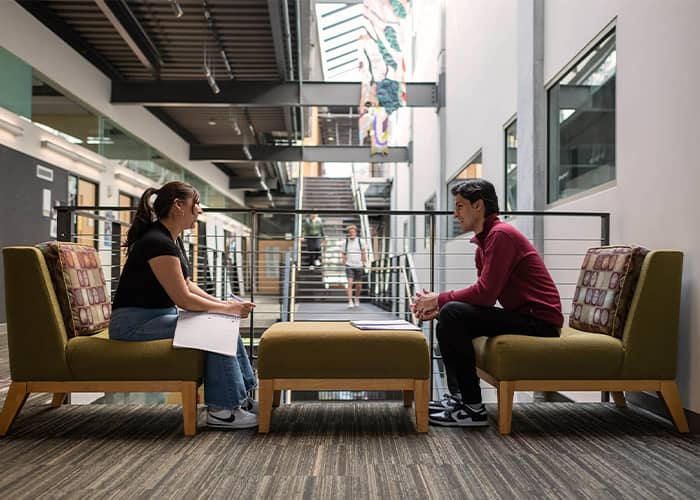 Two students converse, sitting on couches that face each other, in a state-of-the-art academic building.
