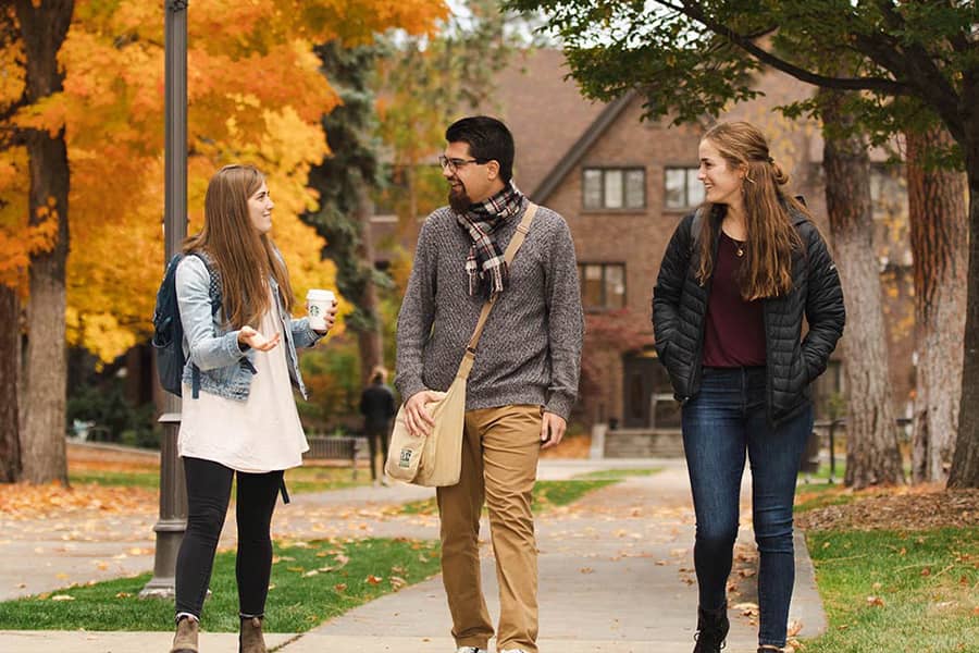 Three students walk along a path past academic buildings on an autumn day.