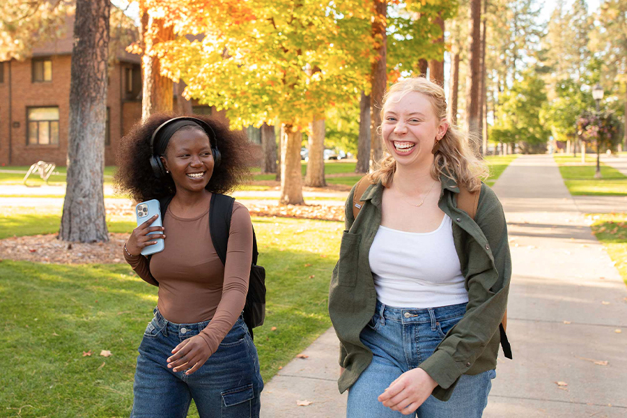 Two students walk and converse outside on a sunny autumn day.