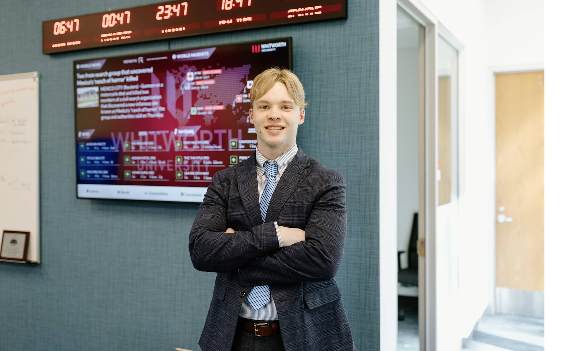 A business student stands, arms crossed, in front of a digital display with stock trading data.