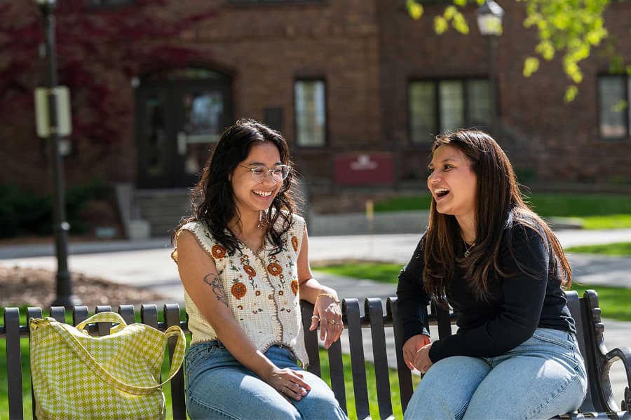 Two students enjoy a lively conversation on a metal bench on a sunny day.