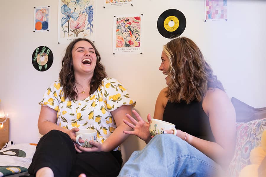 Two students sit and laugh together on a bed in a student dorm room.
