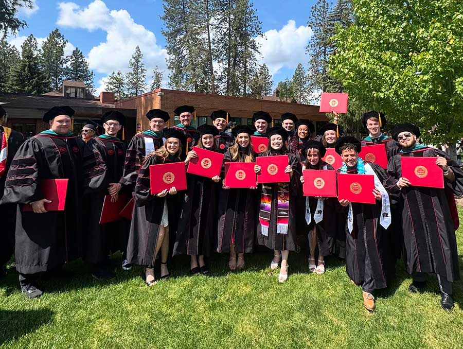 Doctoral graduates in full regalia pose, smiling and raising diplomas, outdoors on a sunny day with trees in the background.