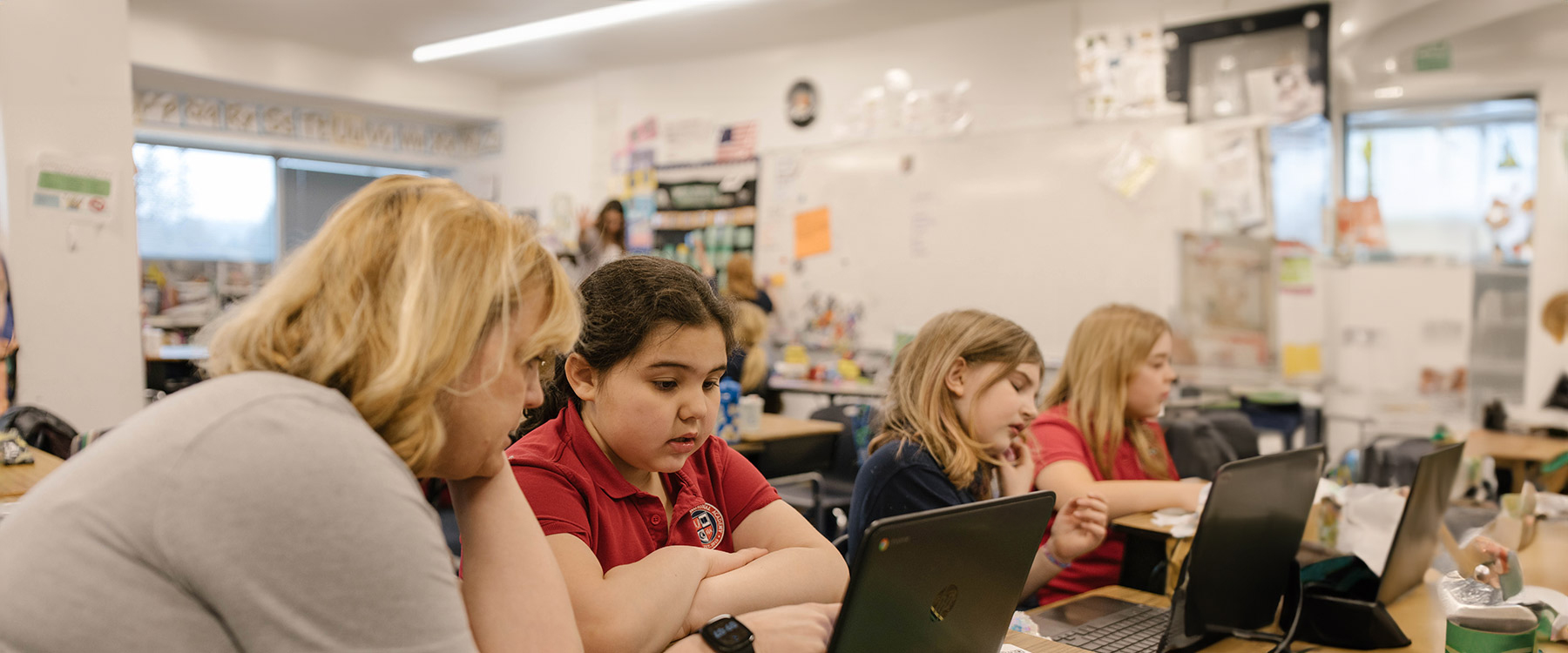A teacher helps a young student with her laptop while other students work at their desks in a classroom setting.