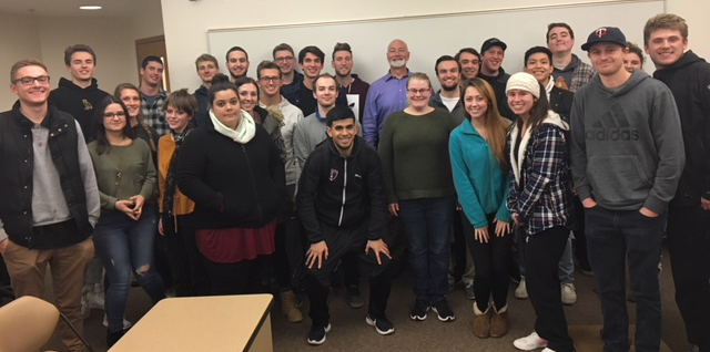 John Perkins and students pose, smiling, in front of a whiteboard in a classroom setting.