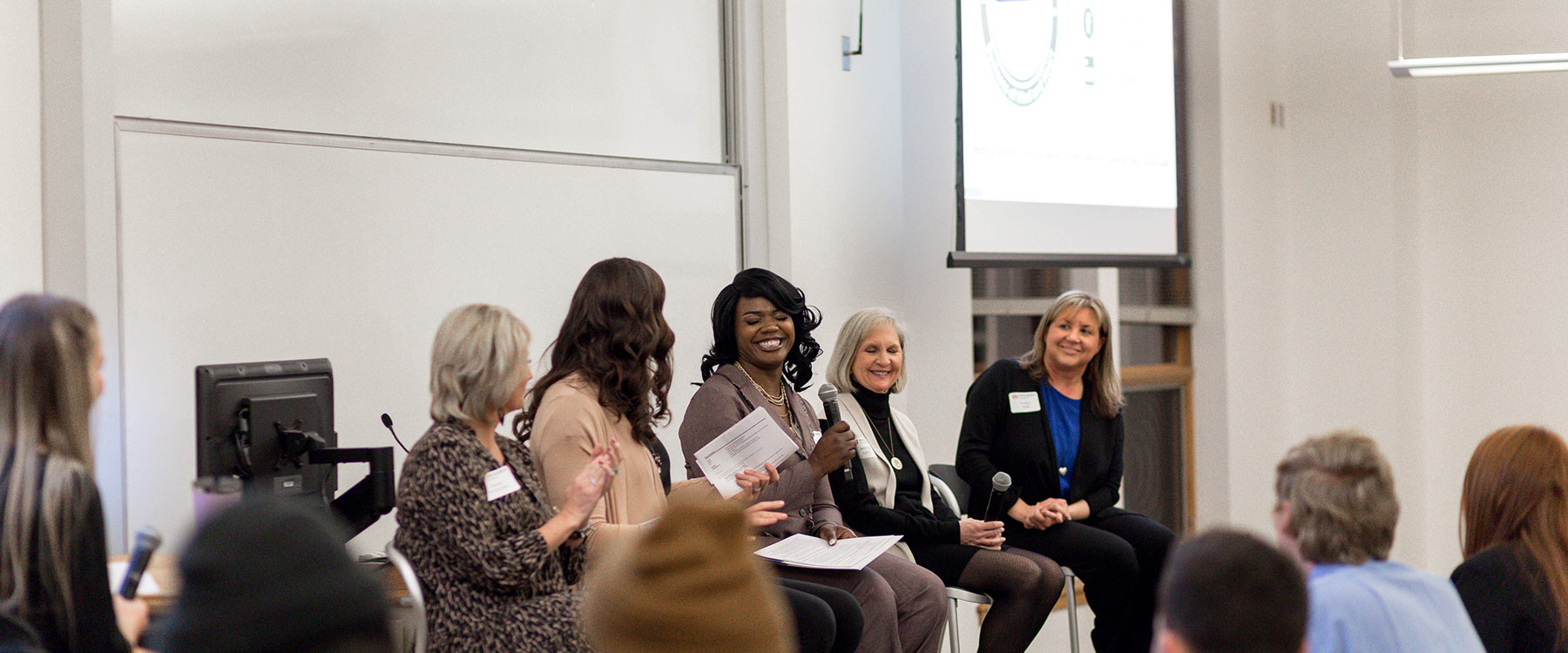 A panel of women laugh as one smiles broadly and talks into a microphone during a presentation in a classroom setting.