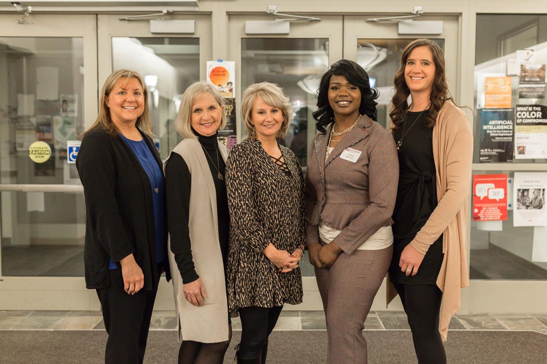 Five professionally dressed women pose together, smiling, in front of glass doors.